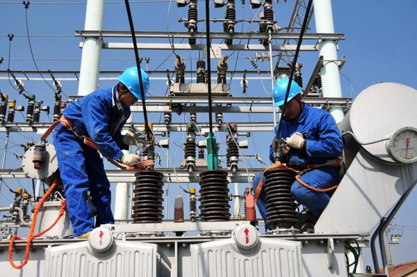 Two technicians wearing blue hard hats work beside the bushings of an outdoor power transformer.