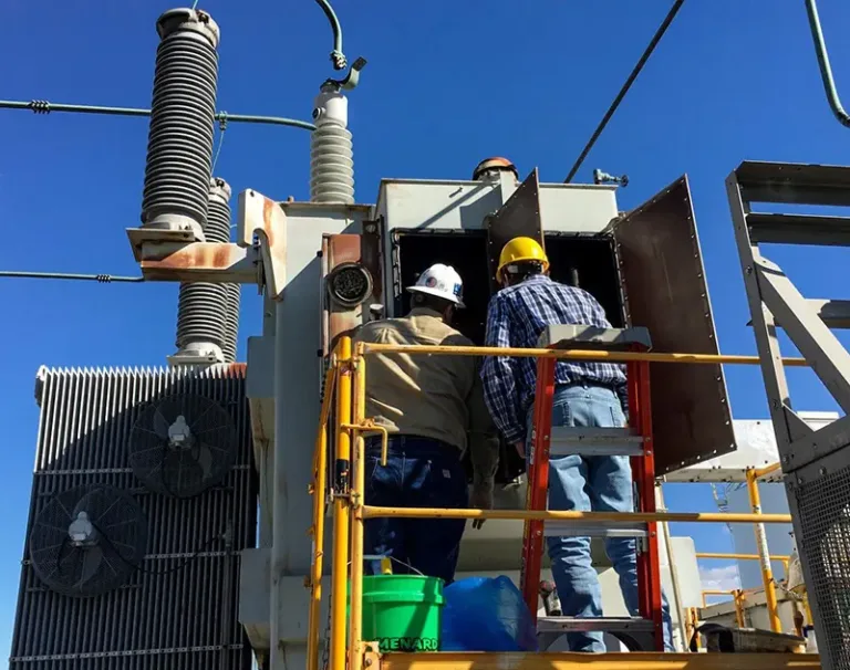 Two technicians inspecting an open cabinet on an outdoor power transformer.