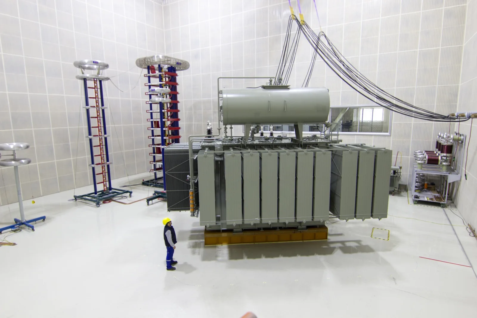 A large power transformer in a high-voltage test hall, with an engineer standing beside it